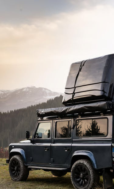Land Rover Defender 110 noir avec une tente de toit rigide garé devant des montagnes enneigées.